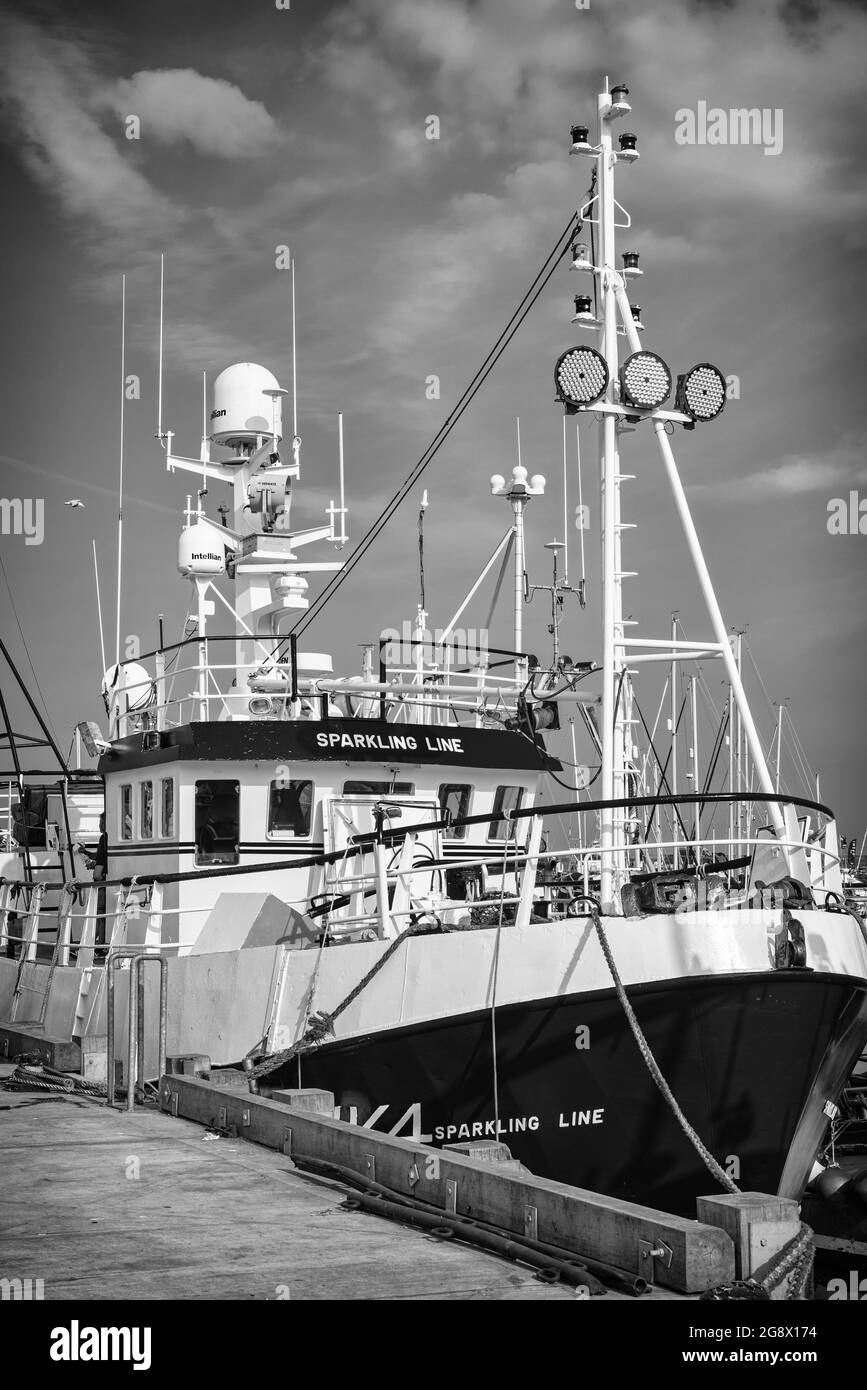 A freshly painted trawler is moored alongside a wharf.  Antennae and radar bristle above the bridge and a sky with clouds is above. Stock Photo