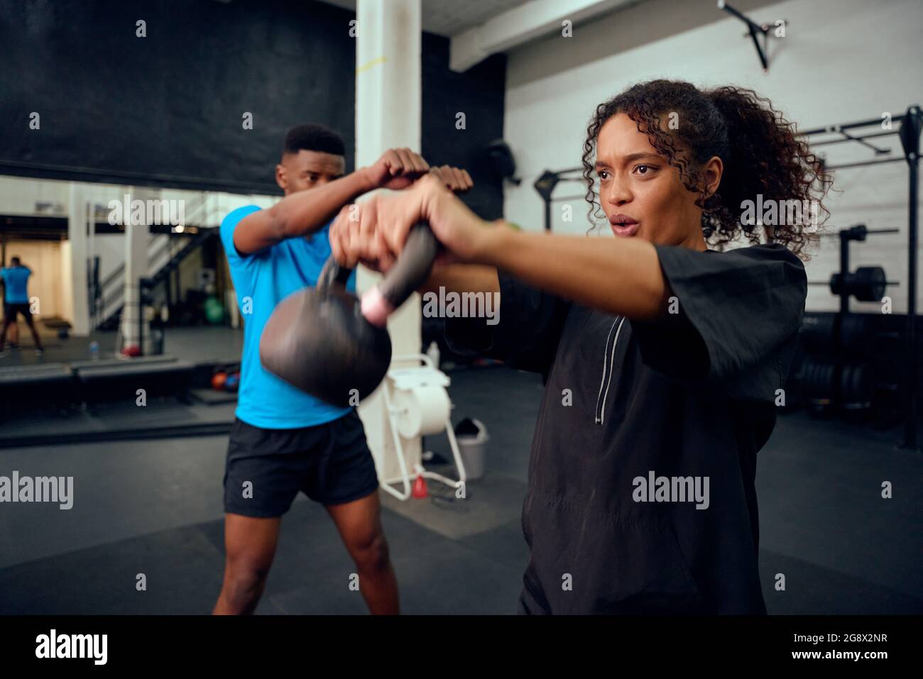 African American male personal trainer instructing African American female with a kettlebell routine in the gym. Mixed race friends doing cross Stock Photo