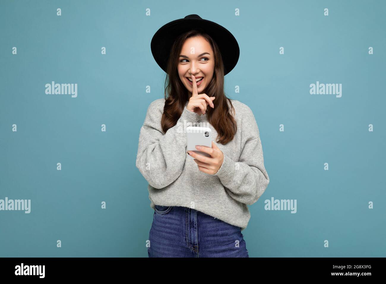 Attractive young smiling woman wearing black hat and grey sweater holding smartphone looking to the side showing shhh gesture isolated on background Stock Photo
