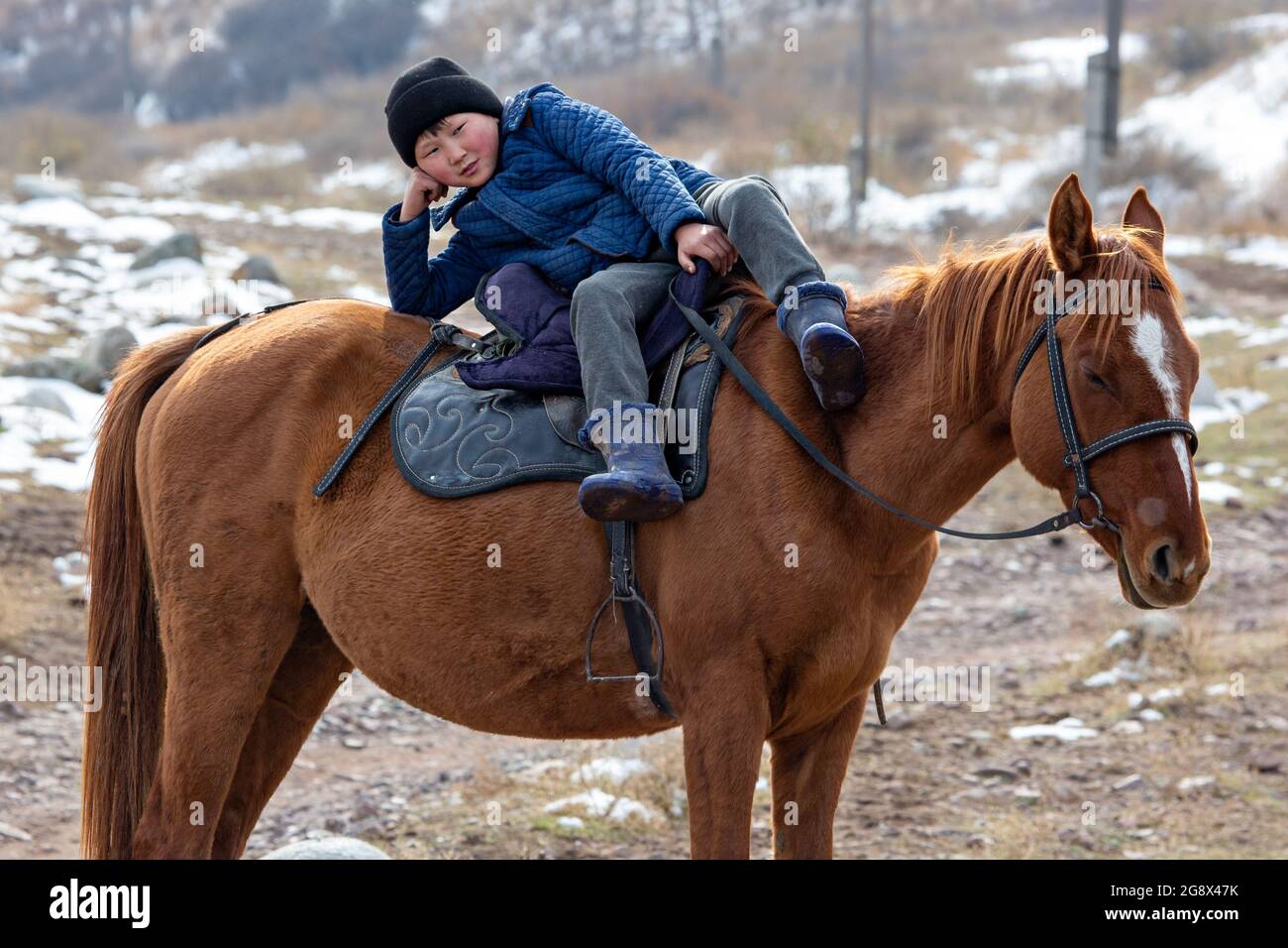 Nomadic young boy lying down over the horse and looks at me, near Bishkek, Kyrgyzstan Stock Photo