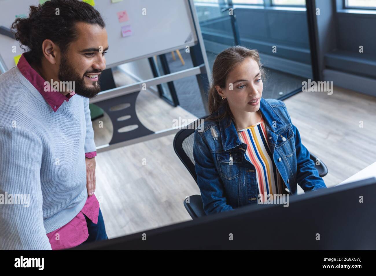 Two diverse creative colleagues sitting at desk using computer and smiling Stock Photo