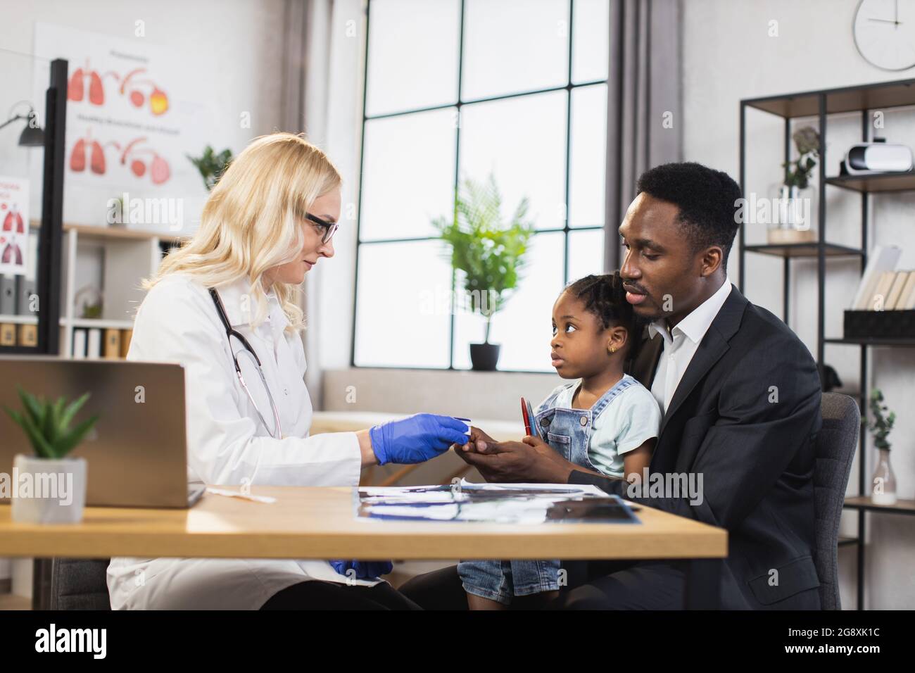 Caucasian doctor checking oxygen saturation of little african girl using modern pulse oximeter. Cute patient sitting on knees af her caring father. Family visiting doctor for controlling health. Stock Photo