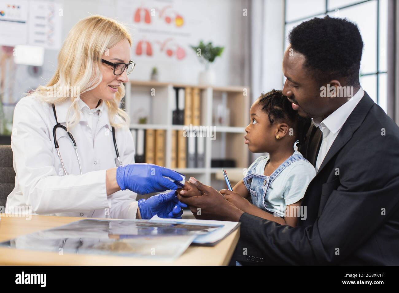 African man keeping his pretty daughter while caucasian pediatrician using pulse oximeter for checking saturation. Concept of people, medicine and health control. Stock Photo