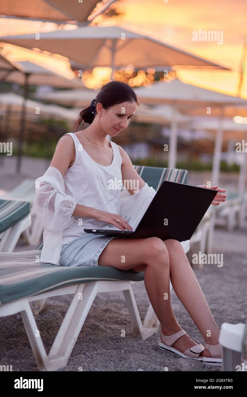 Young brunette with a laptop on a sunbed on the beach. Freelancer, working remotely, webinar, learning concept, chatting with friends. New normal concept Stock Photo