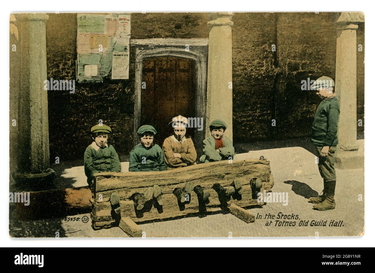 Early 1900's tinted greetings card of young boys sitting in the stocks, an antiquated form of punishment through humiliation of the criminal, though posed for this photo. Totnes Old Guild Hall, Devon, U.K. posted 1913 Stock Photo