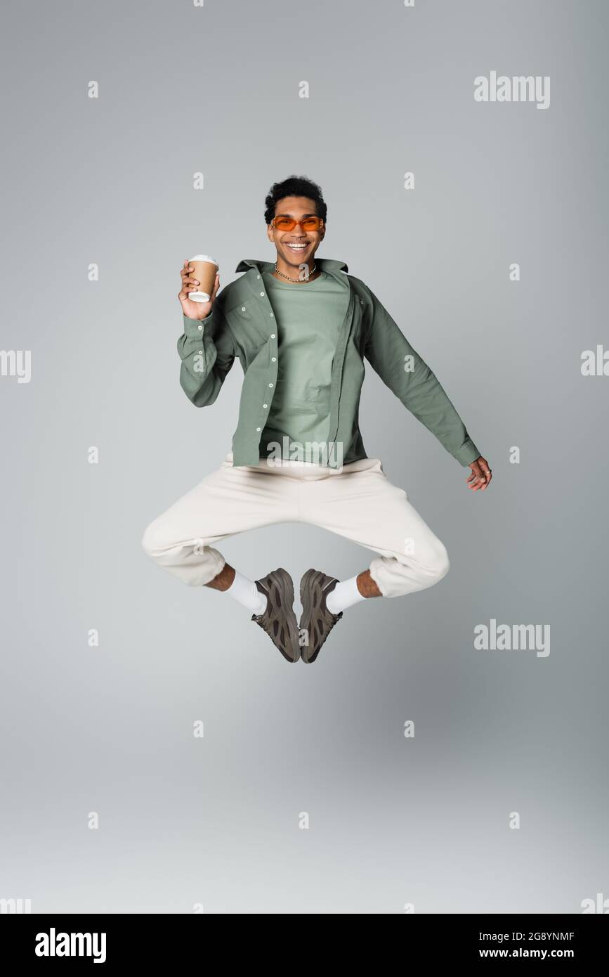 excited african american man in trendy clothes jumping with paper cup isolated on grey Stock Photo