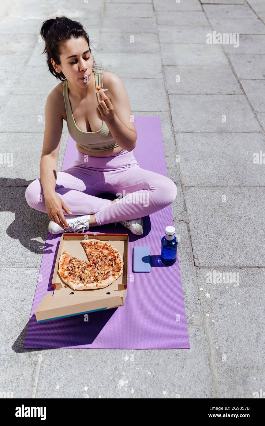 Female athlete eating pizza while sitting cross-legged on exercise mat Stock Photo