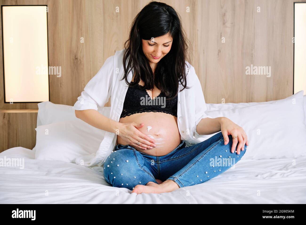 Pregnant woman applying moisturizer on belly while sitting on bed in bedroom Stock Photo