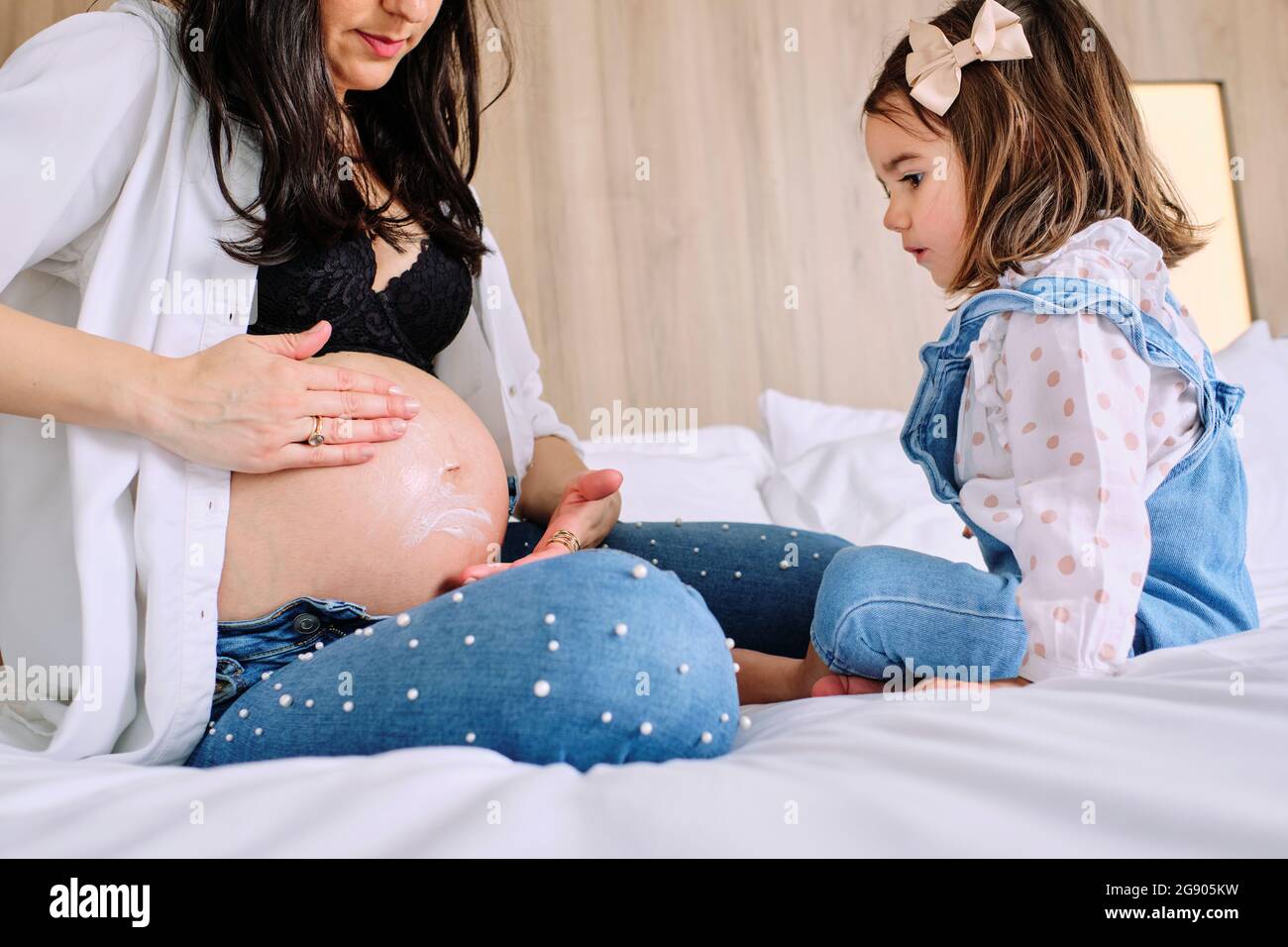 Girl looking at mother applying moisturizer on belly while sitting on bed Stock Photo