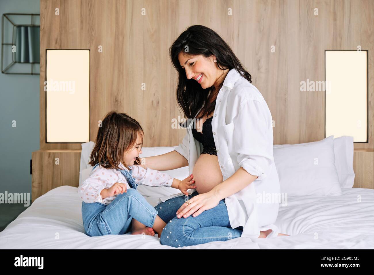 Cute girl touching pregnant mothers belly while sitting on bed at home Stock Photo