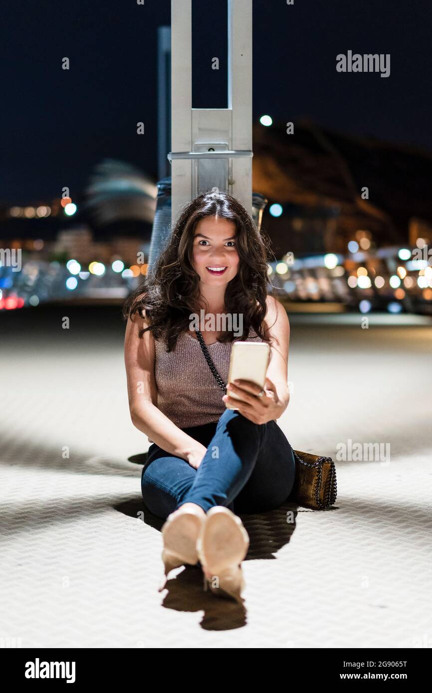 Smiling woman with mobile phone sitting on floor at pier during night Stock Photo