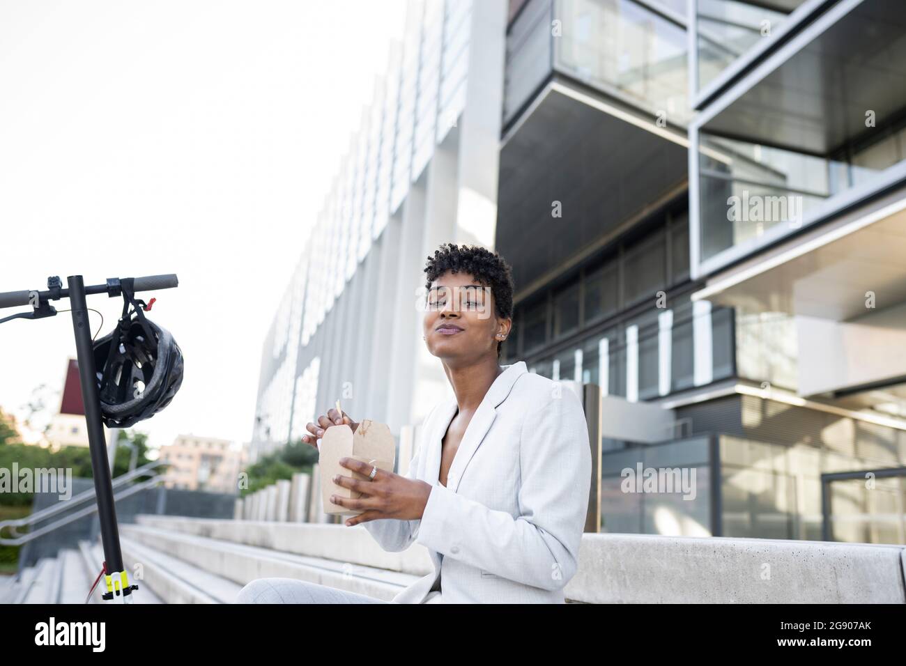 Businesswoman eating meal while sitting with electric scooter on steps Stock Photo