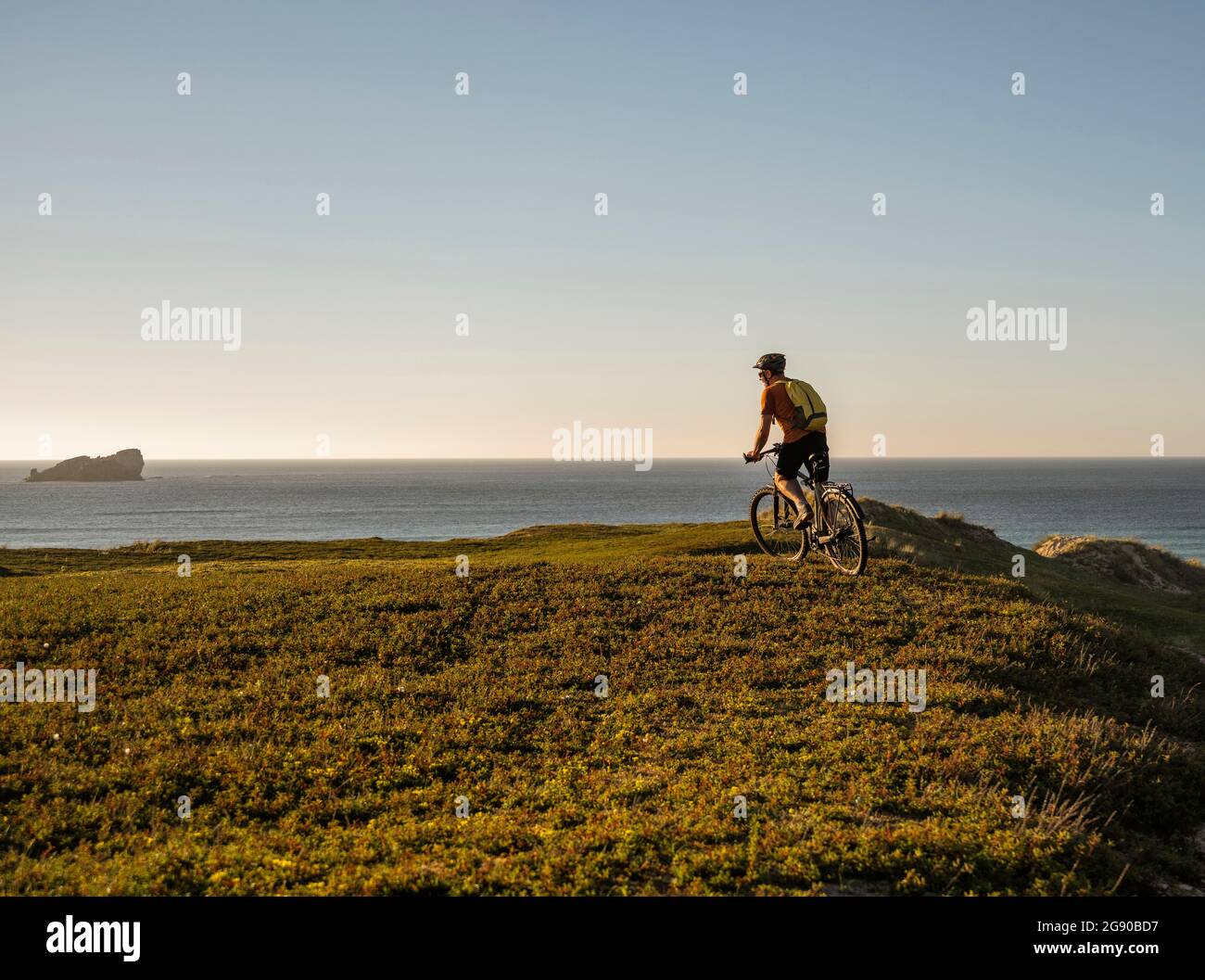 Male athlete with backpack cycling on green grass at sunset Stock Photo