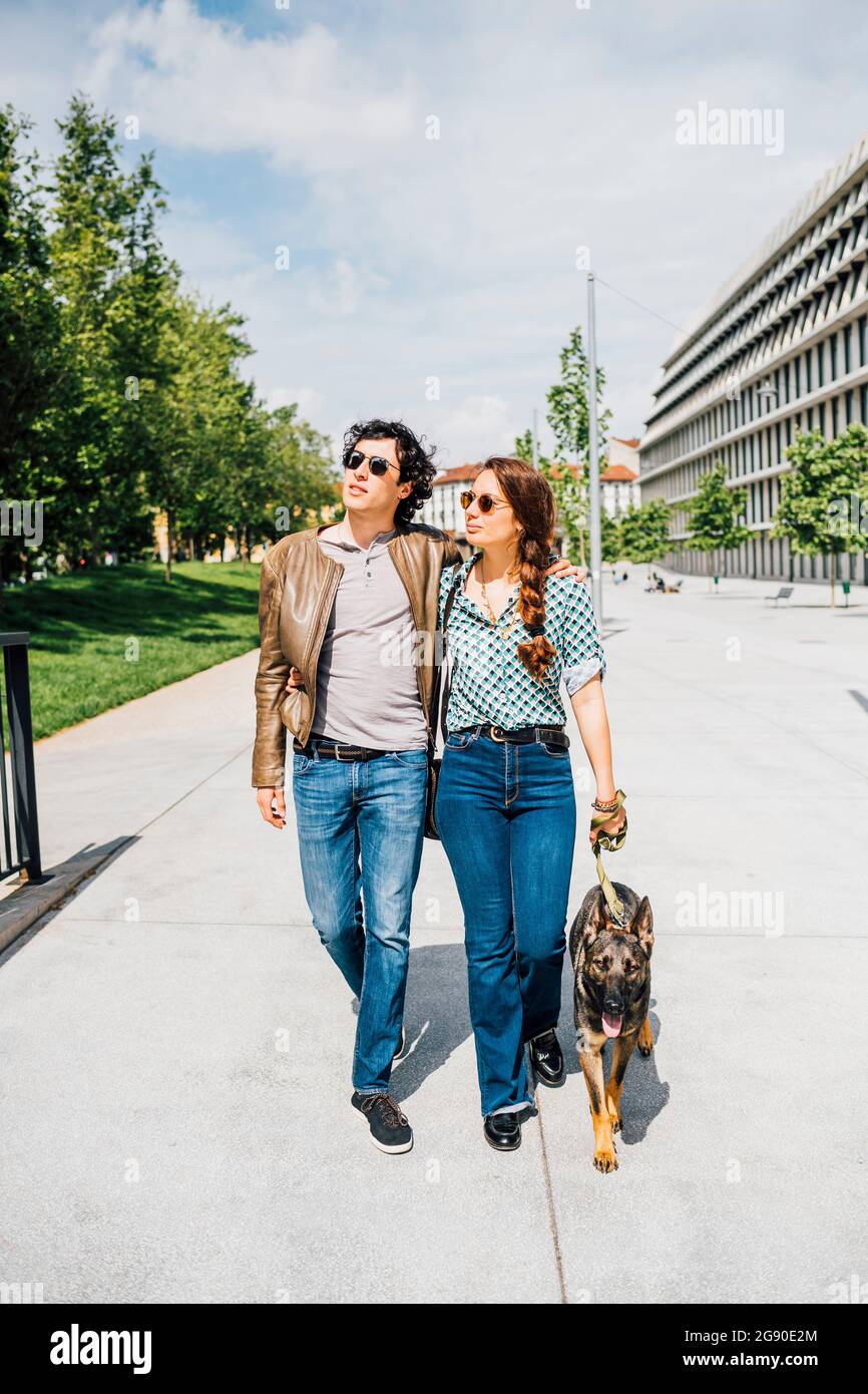 Couple walking with dog on city street during sunny day Stock Photo