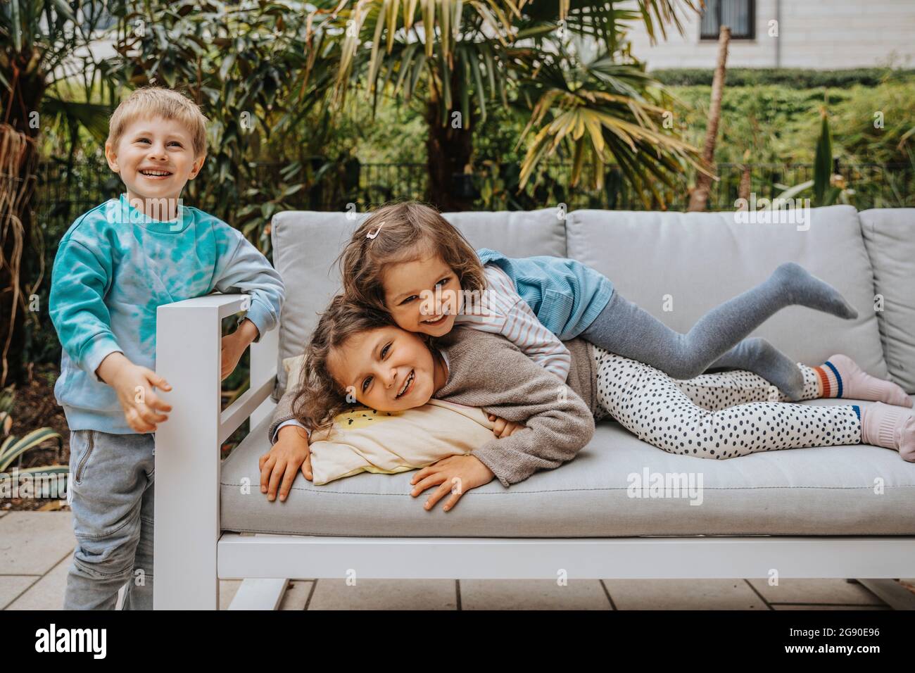 Boy standing by sofa while sisters enjoying at backyard Stock Photo