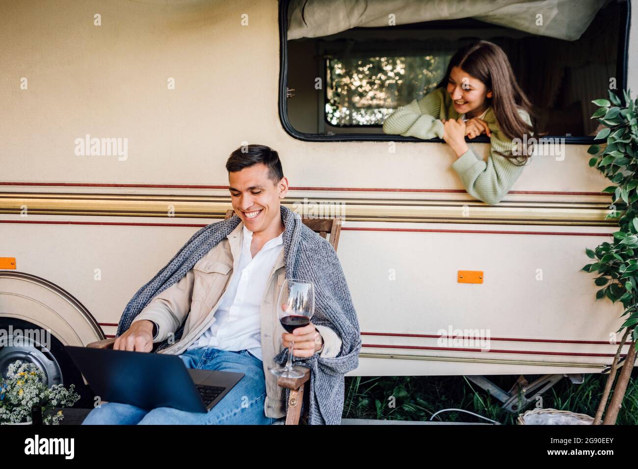 Happy woman looking through camper trailer window at boyfriend using laptop Stock Photo