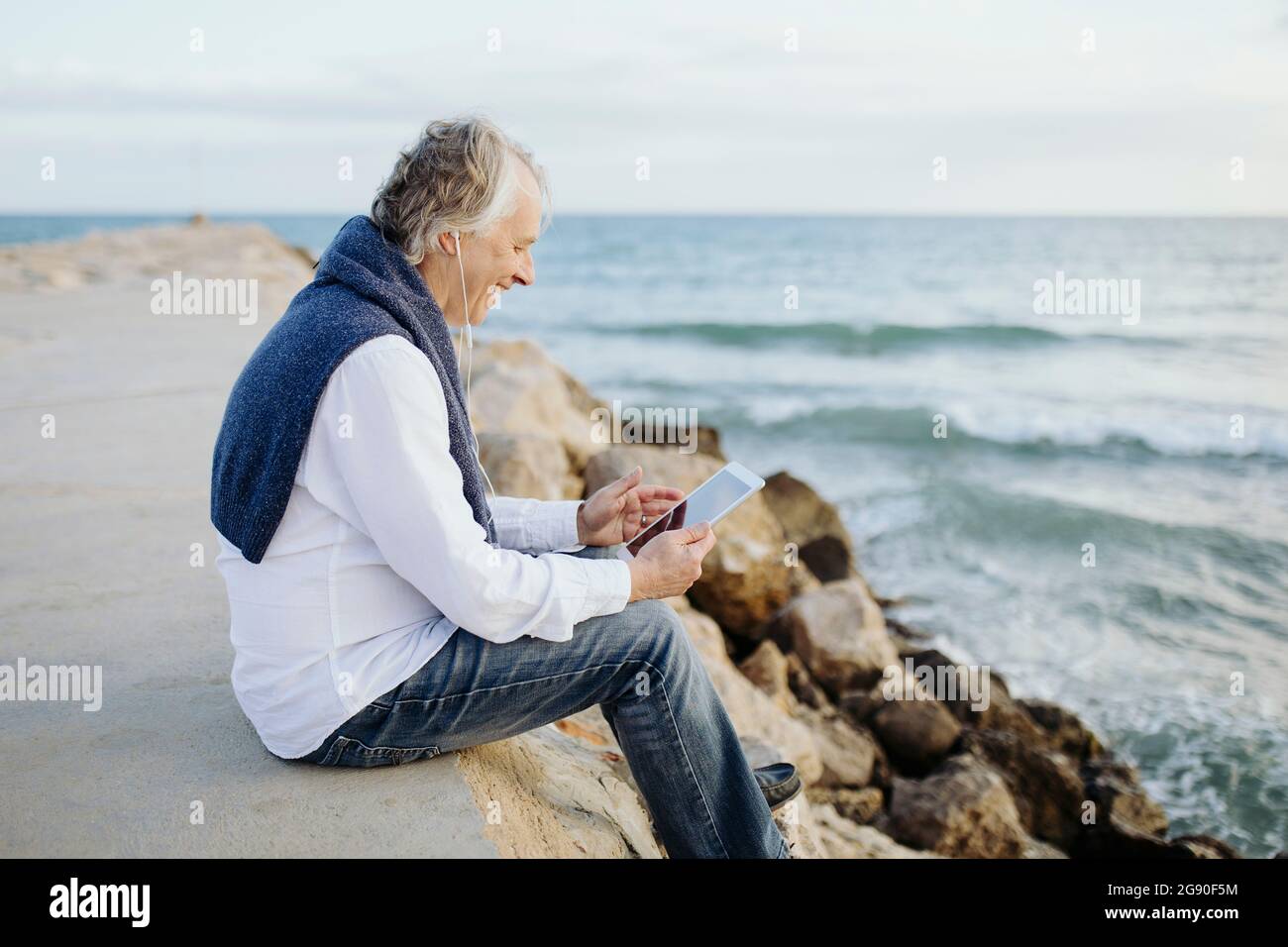 Happy mature man using digital tablet sitting at seaside Stock Photo