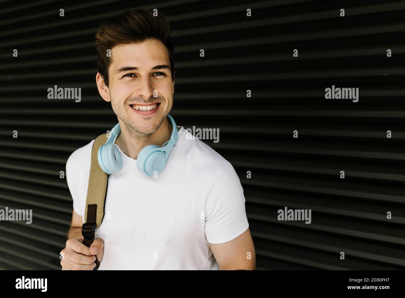 Smiling young man looking away while carrying backpack in front of shutter Stock Photo