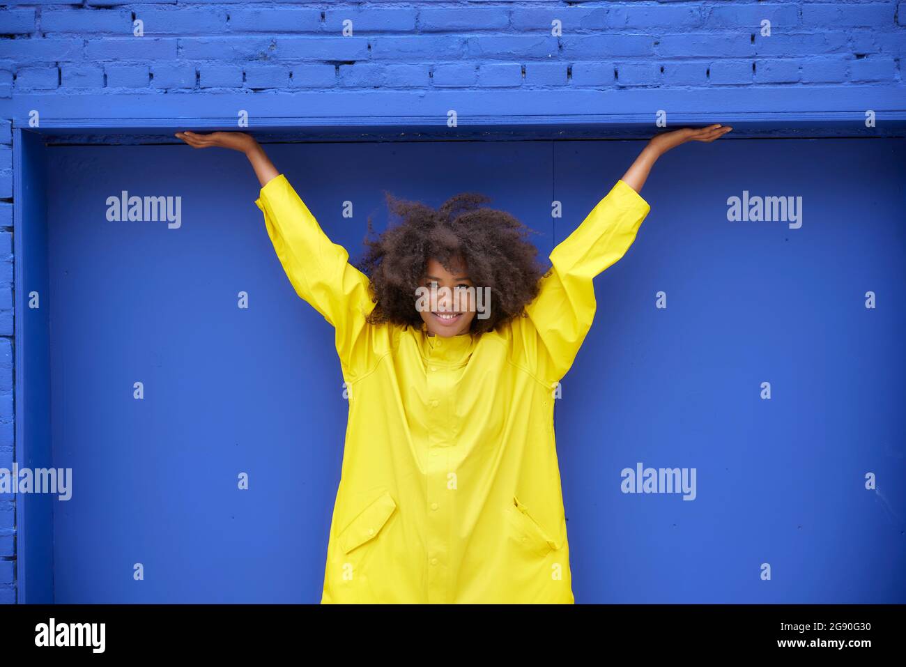 Smiling young Afro woman with arms raised in front of blue wall Stock ...