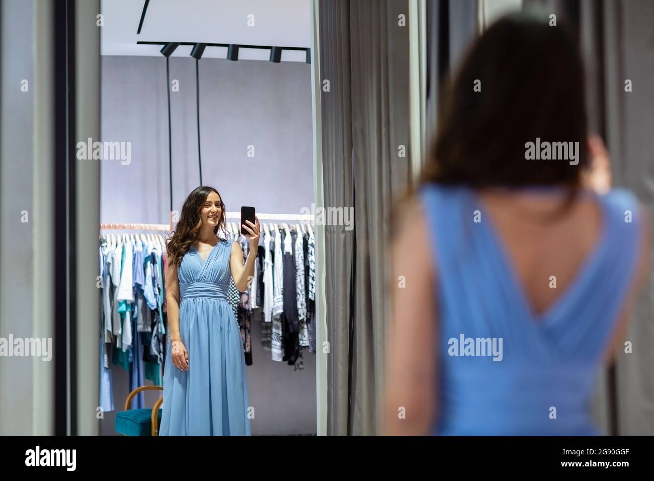 Young woman photographing self in mirror wearing blue dress at boutique Stock Photo