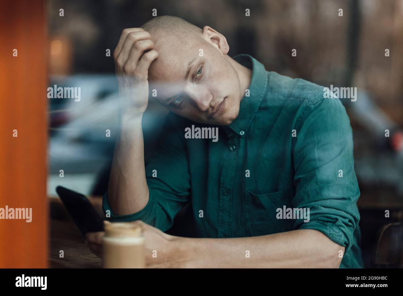 Businessman holding mobile phone while scratching head seen from cafe window Stock Photo