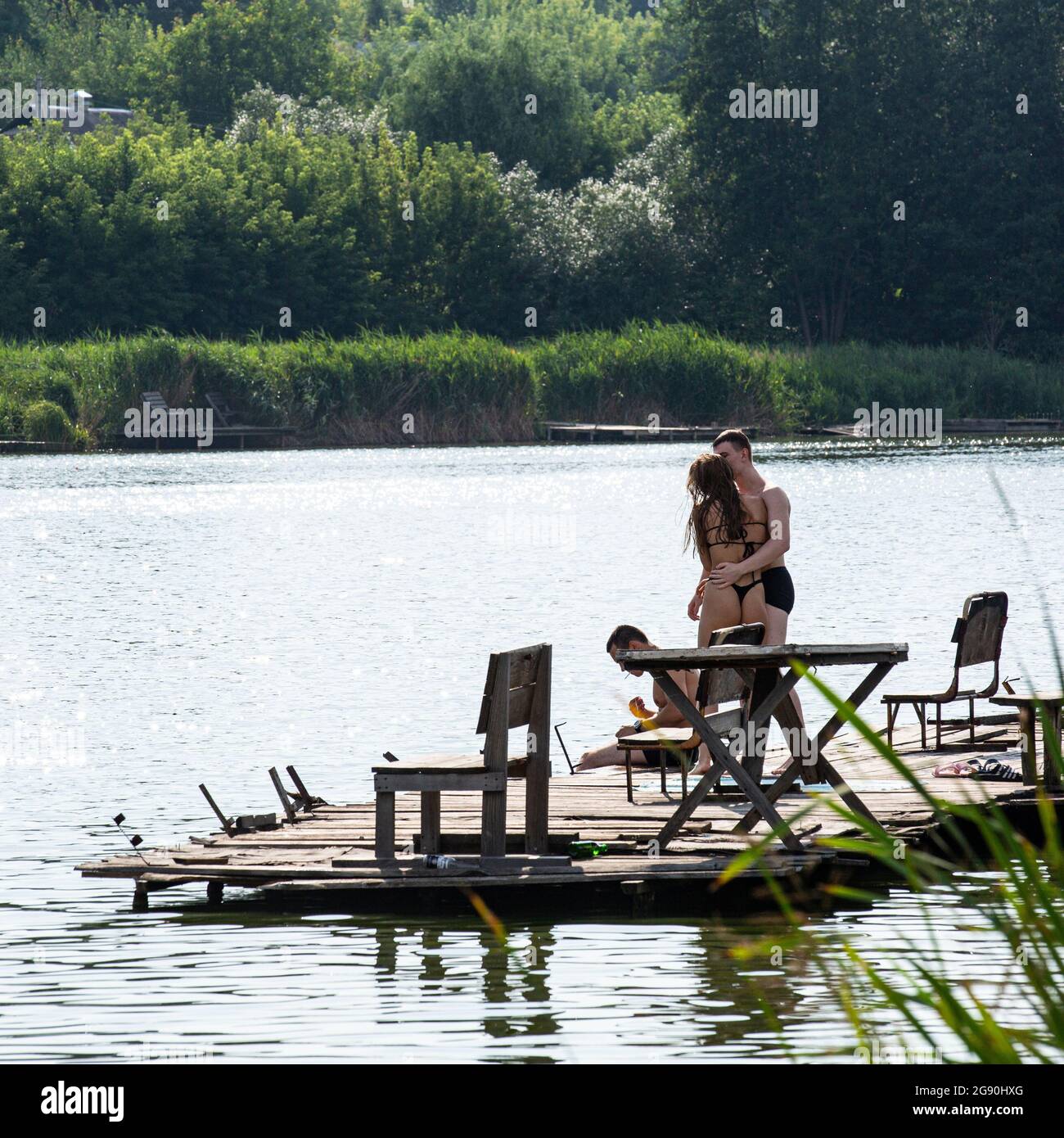 Young couple hugs on the wooden pier on lake in Muzychi in Kiev region, Ukraine on sunny July day their friend sits on the pier his legs in the water. Stock Photo