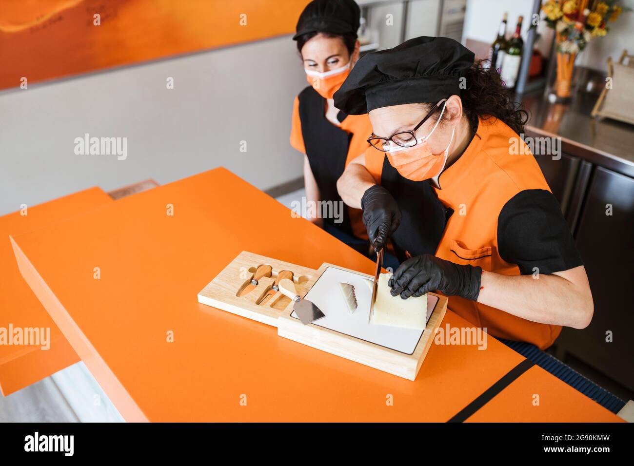 Waitress looking at female chef cutting cheese on board during COVID-19 Stock Photo