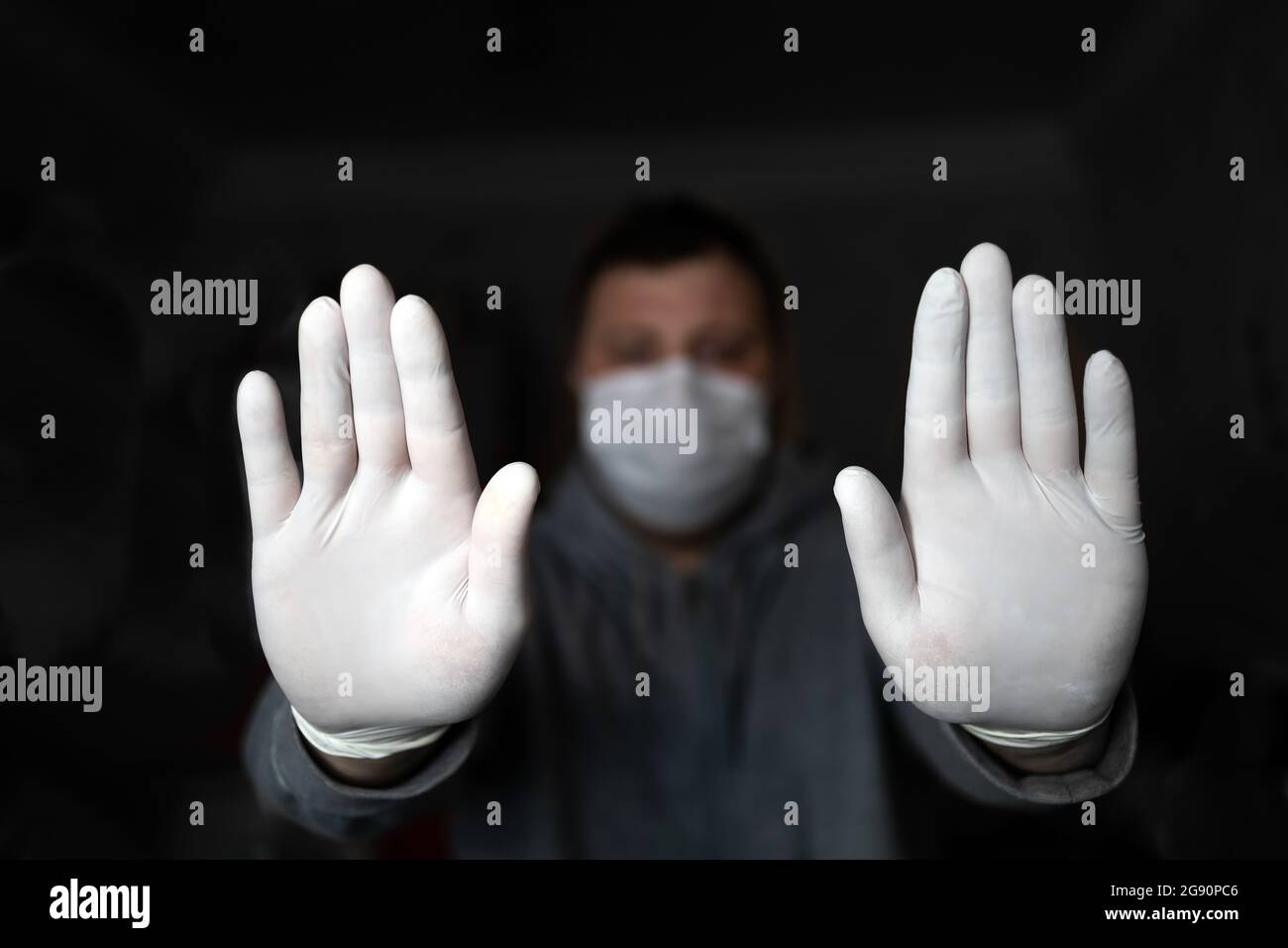 A man in a medical mask and gloves shows STOP with two hands, on a dark background. Coronavirus infection protection concept. COVID-19. Stock Photo