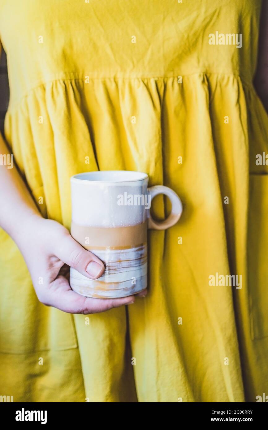 Cropped young white woman in a simple yellow linen dress holds in her hands a clay ceramic cup of an original handmade shape. Copy space. Trend. Stock Photo