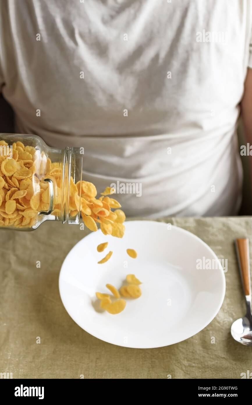 A cropped man sprinkles cornflakes in a white plate on a plain rough tablecloth. Close-up. Top view. Selective focus. Concept, simple fast american he Stock Photo
