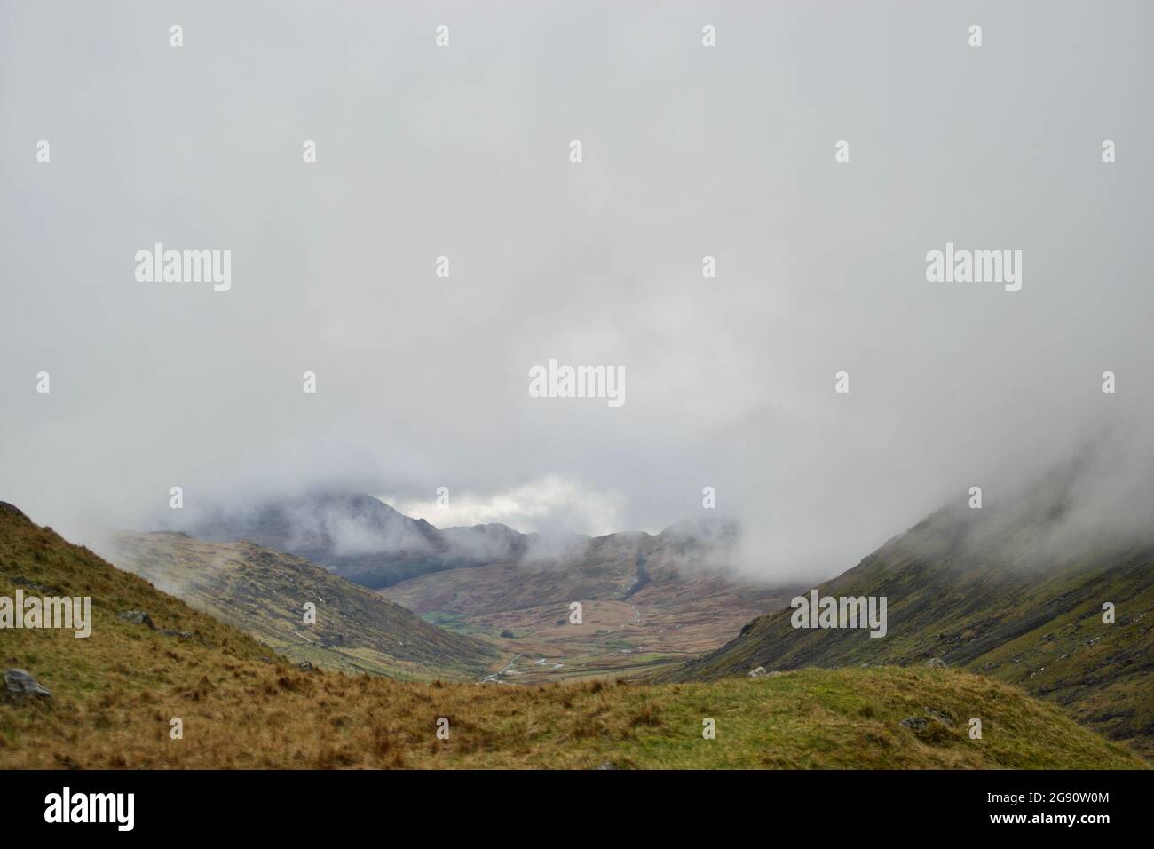 A misty mountain valley: clouds hanging low around mountain peaks; grass in foreground; a valley with a road snaking down - bends in the route across Stock Photo