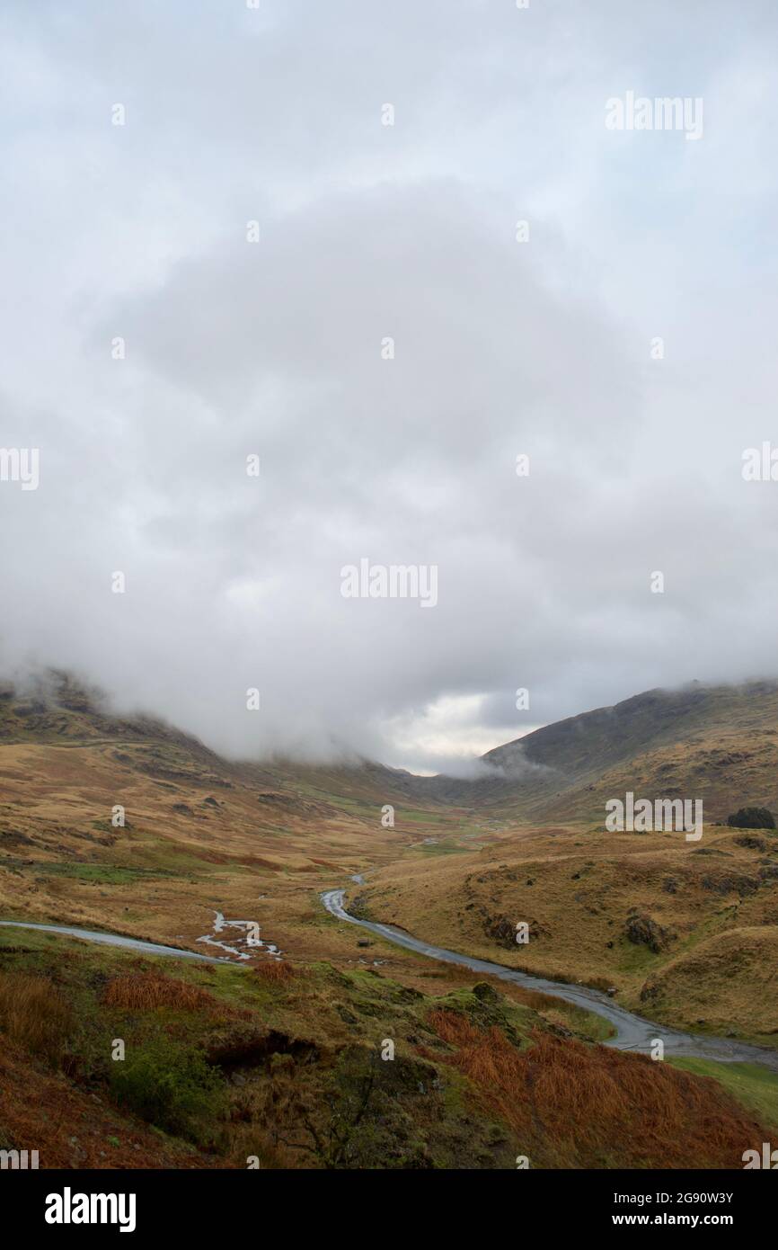 A mountain road (pass): single track road snaking between brown grassy moorland, the mountain peaks hidden in low cloud and mist. Autumn colours in wi Stock Photo
