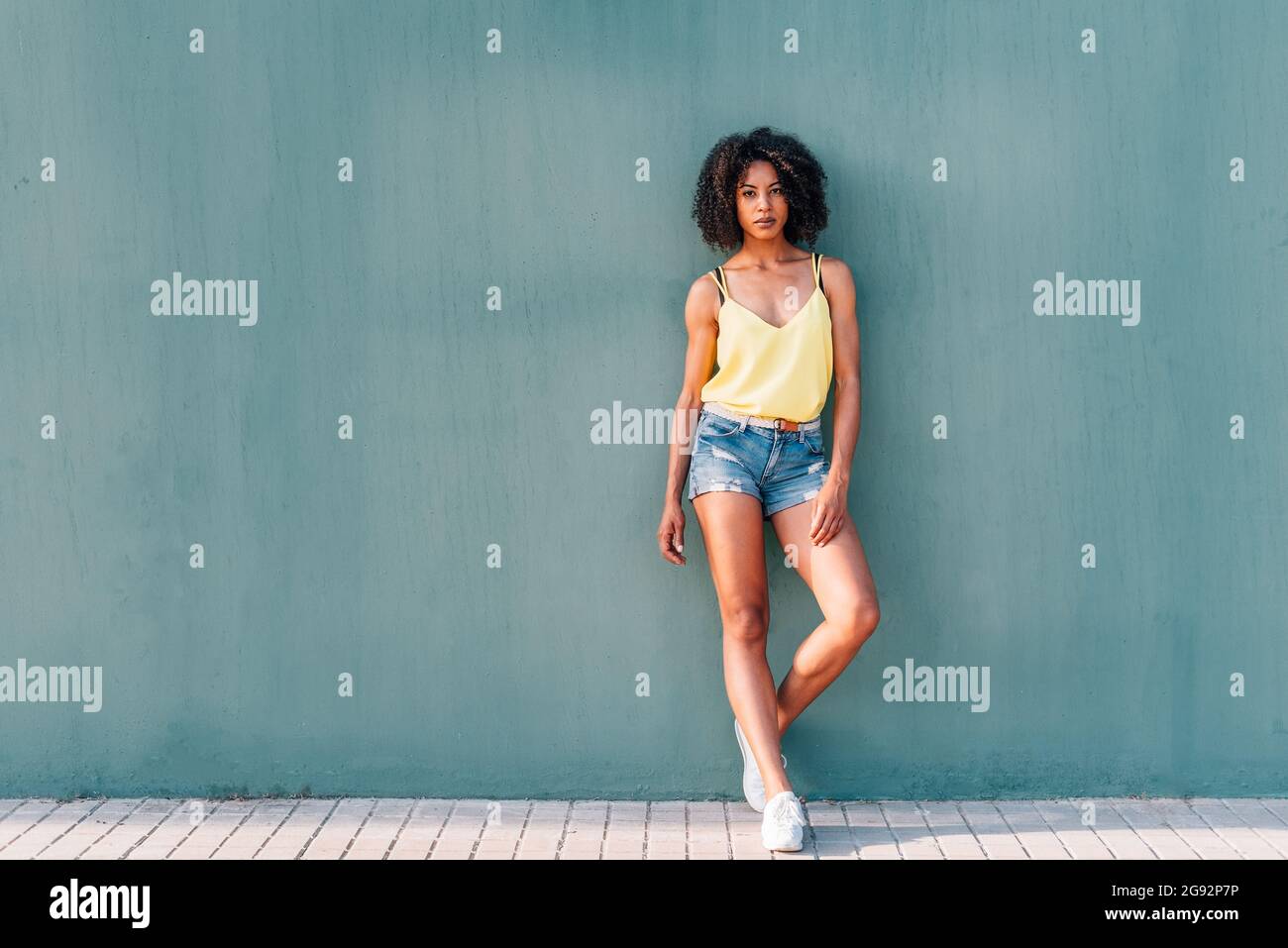 horizontal portrait of an african woman with a green wall background. She is leaning on green wall with summer clothes and looking at camera Stock Photo