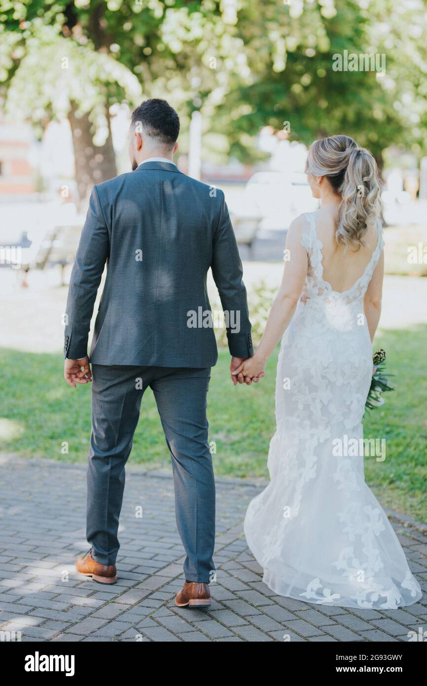 Back view of a stylish wedding couple walking in the park holding hands ...