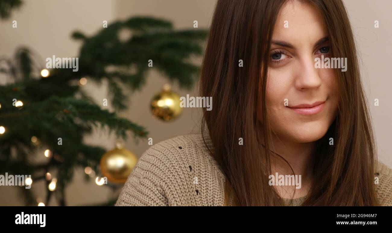 Shy young woman is looking at camera. She is smiling. Camera is bring nearer to her face. Behind her is standing christmas tree. Stock Photo