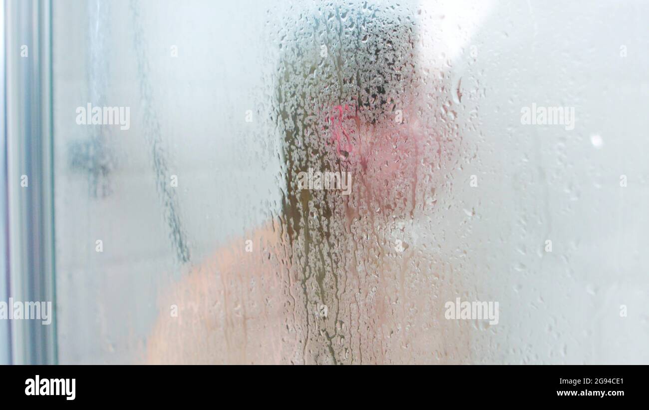 Unrecognizable woman standing in shower cabin behind steamy glass with waterdrop. Stock Photo