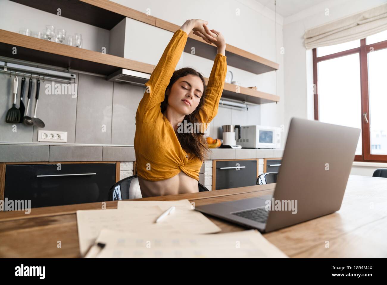 Smiling young brunette woman working on laptop computer while sitting in the kitchen, stretching hands Stock Photo