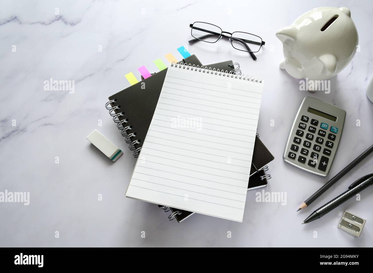 Office desk with a blank notebook, piggy bank, calculator and stationary supplies on a marble surface, copy space, selected focus, narrow depth of fie Stock Photo