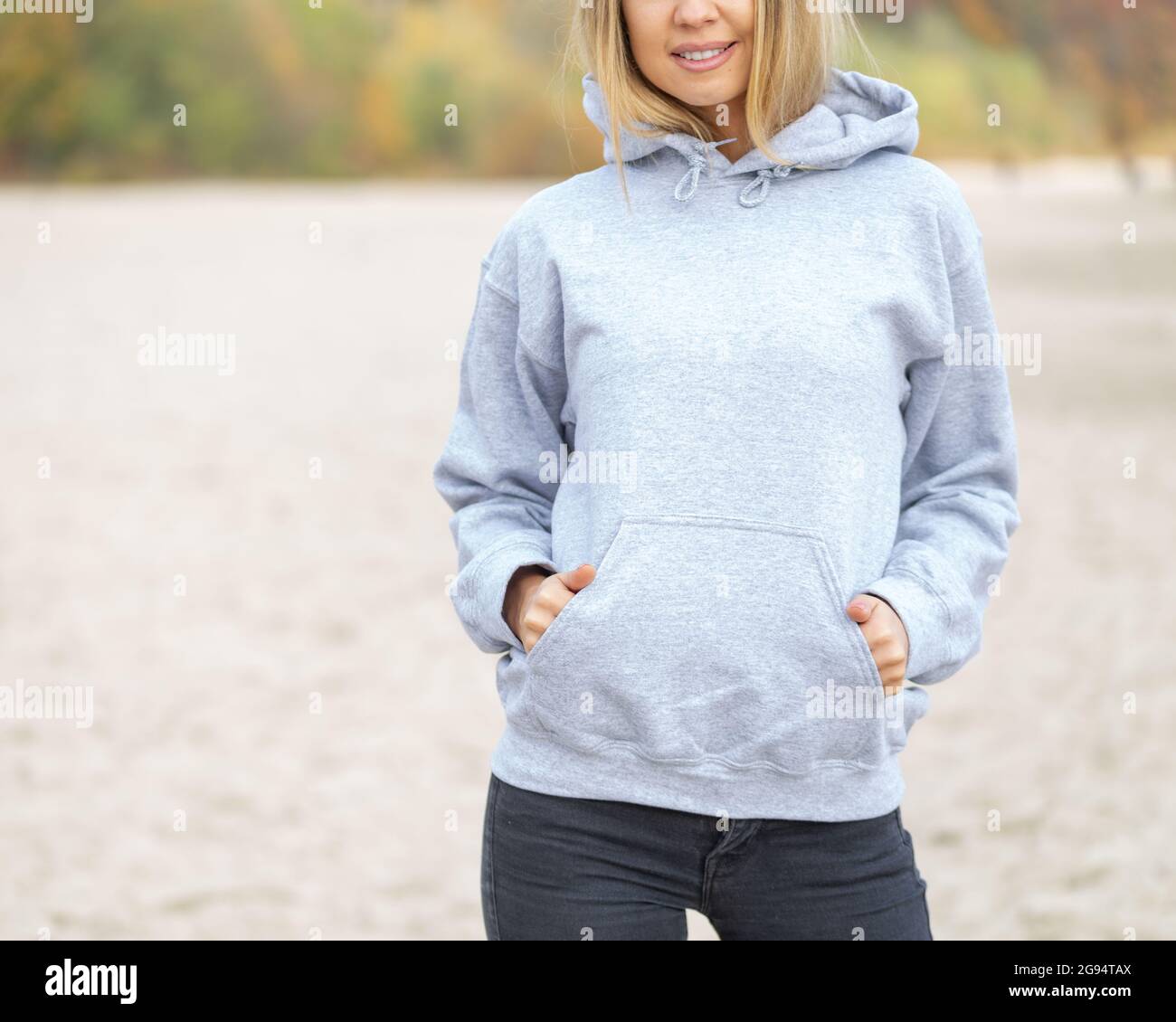 The front view of unrecognizable woman wearing light grey hoodie. She stands on the beach. Copy space on empty area on her blouse for design or inscription. Fashion mockup. Stock Photo