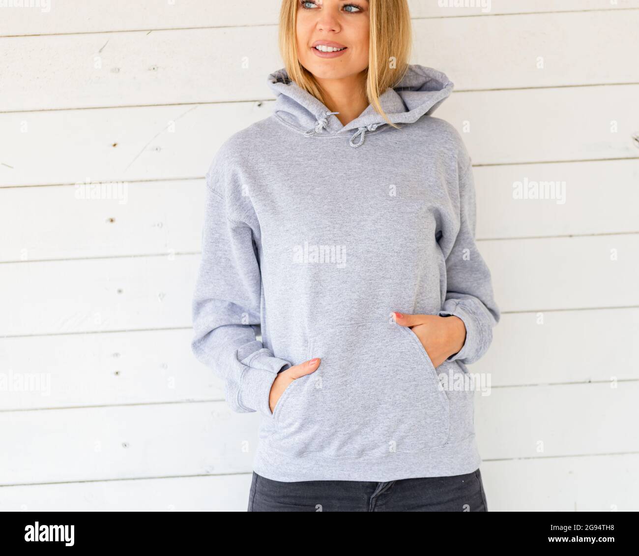 Smiling blond woman wears light grey hoodie. She stands in front of camera, behind her is white board. There is empty area on her hoodie for inscription, clothing design for fashion mockup. Stock Photo