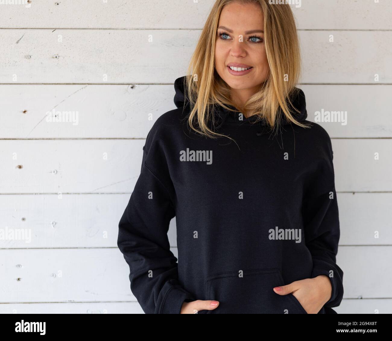 A smiling blonde is standing in a black sweatshirt on a background of white boards, in front of camera. There is a blank space on the garment for a design, logo or inscription. Mockup for clothes. Stock Photo