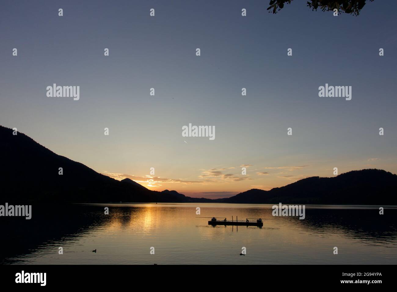 Yellow golden sun setting over beautiful lake Fuschl as ducks have last swim of the day in golden water reflection with pontoon & mountain silhouette Stock Photo