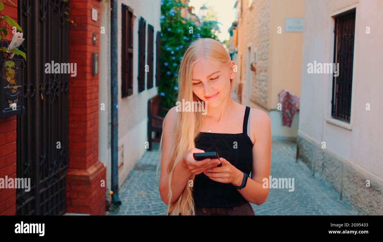 Pretty blonde girl chatting on smartphone while walking down the street of the old town. Smiling and happy woman. Stock Photo