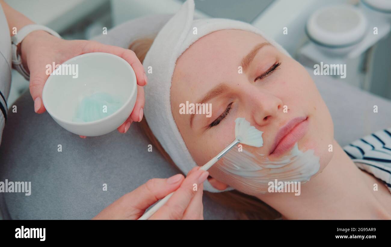 Cosmetician hands applying cream mask on young woman's face at beauty spa salon. Facial skin care treatments. Stock Photo