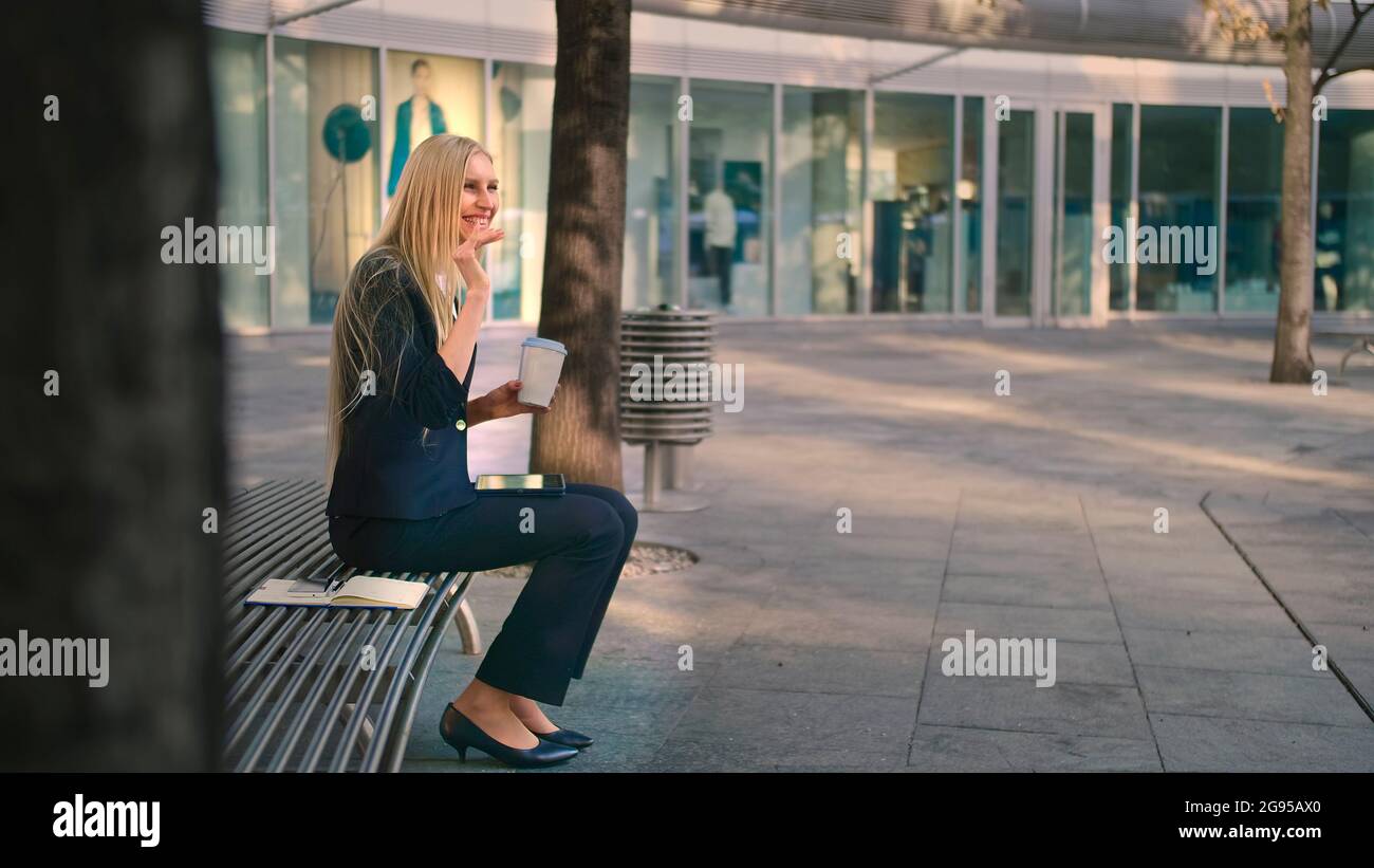 Side view of formal woman sitting with tablet on bench and drinking coffee looking pensively away in leisure Stock Photo