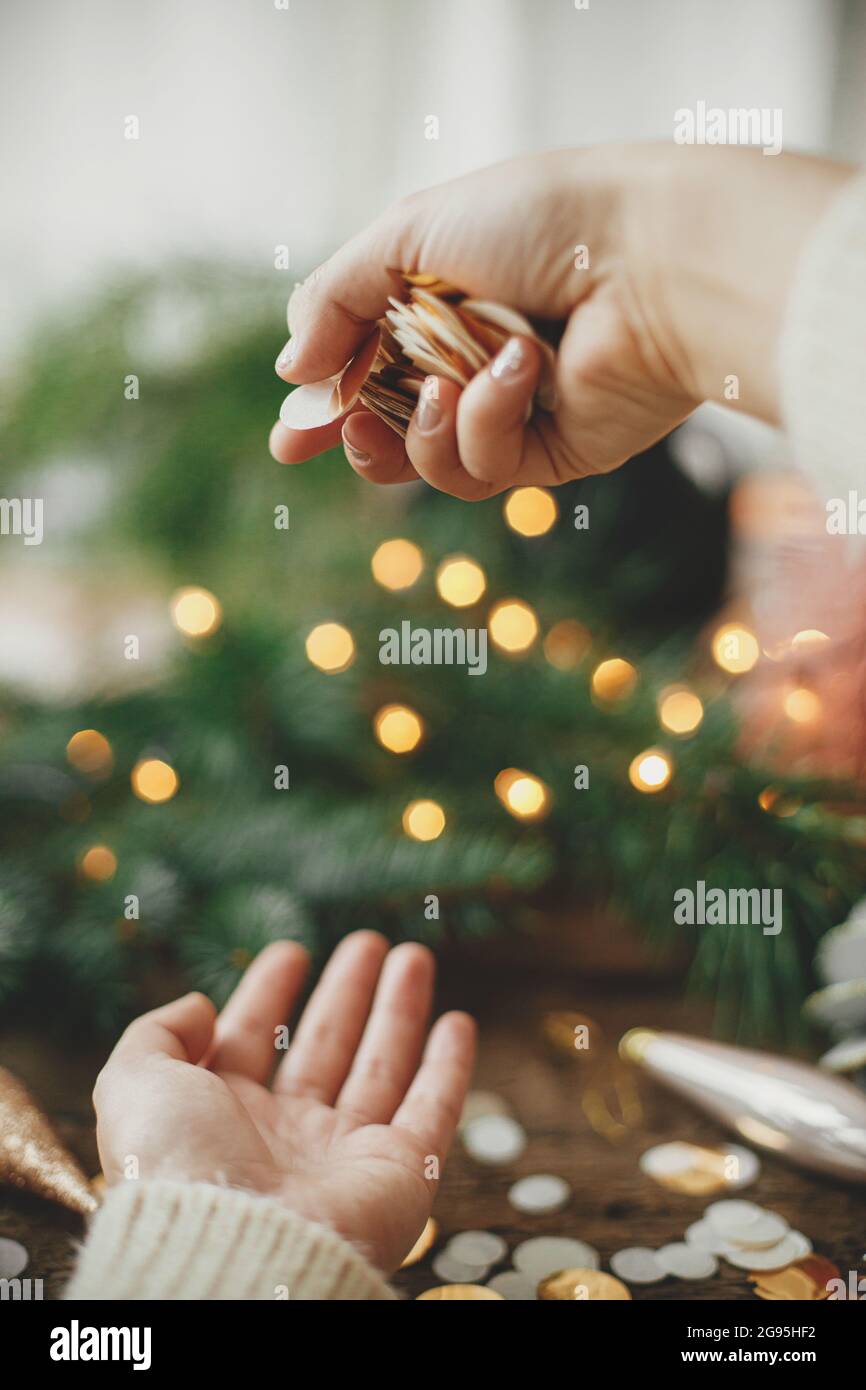 Hands in sweater holding golden confetti on background of christmas lights, decorations, baubles and pine branches on rustic wood. Festive mood. Space Stock Photo