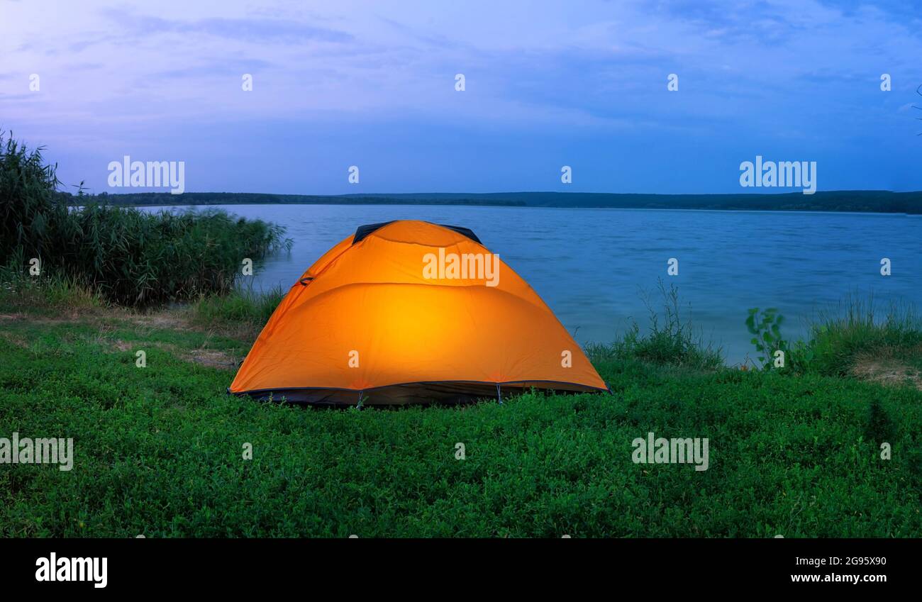 Orange tent illuminated from the inside by lake at dusk Stock Photo