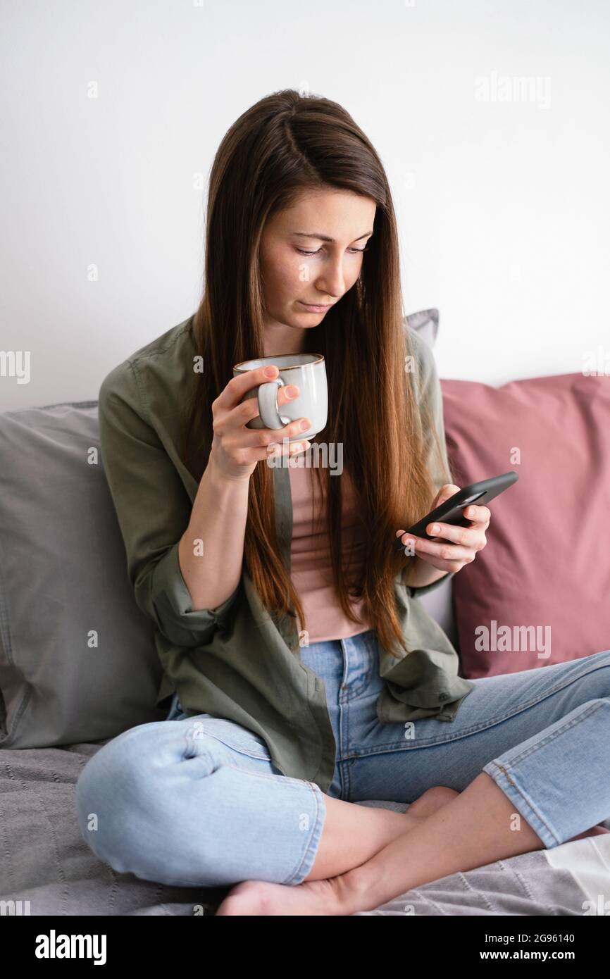 Young woman sitting in bed with cup of tea and using smartphone. Home life. Slow life concept. Stock Photo