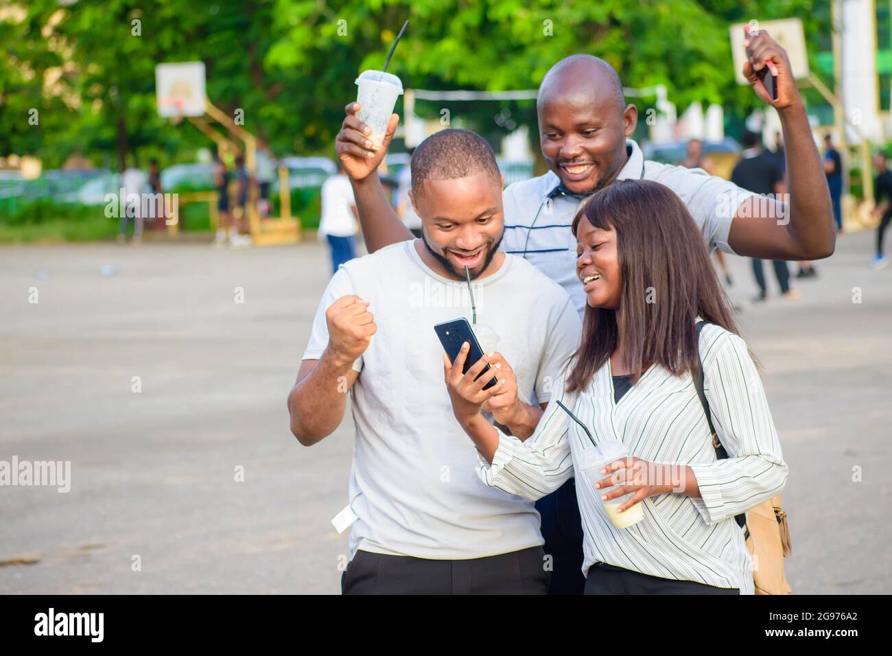 happy group of African friends consisting of two guys and a lady joyfully looking at a smart phone in an outdoor park while jubilating Stock Photo