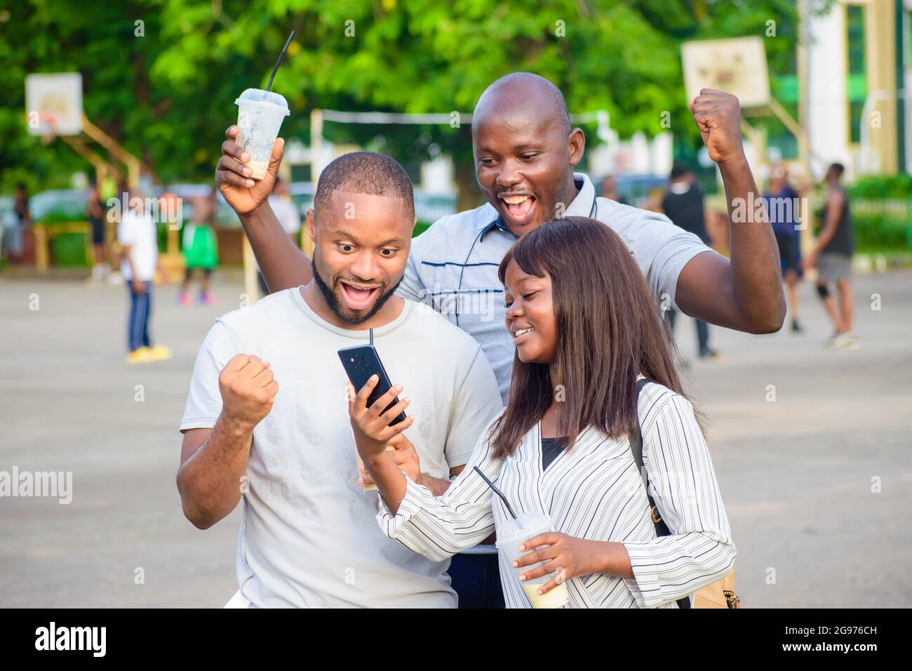 happy group of African friends consisting of two guys and a lady joyfully looking at a smart phone in an outdoor park while jubilating Stock Photo